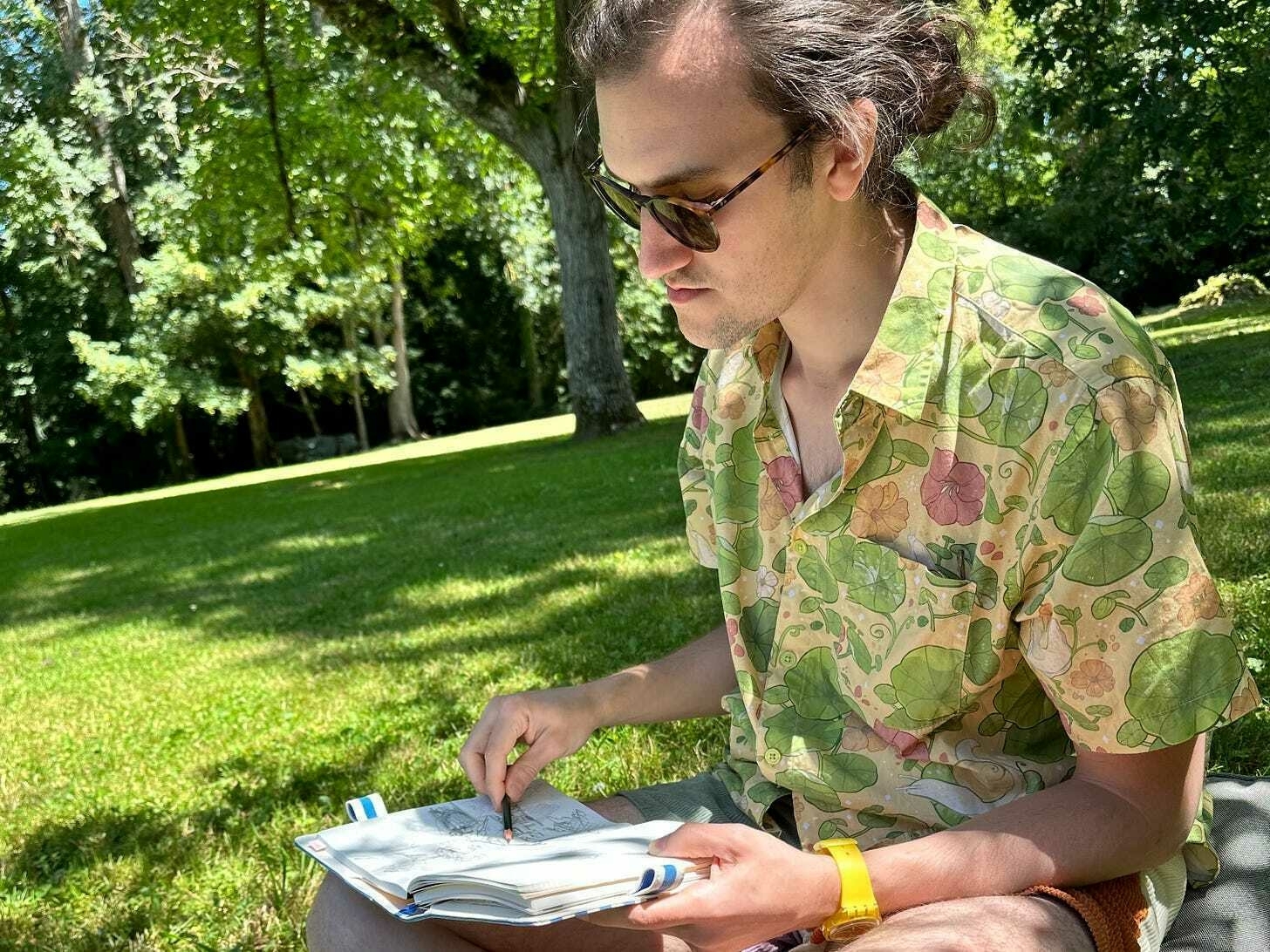 A photo of Simon sitting in the grass drawing in a notebook.